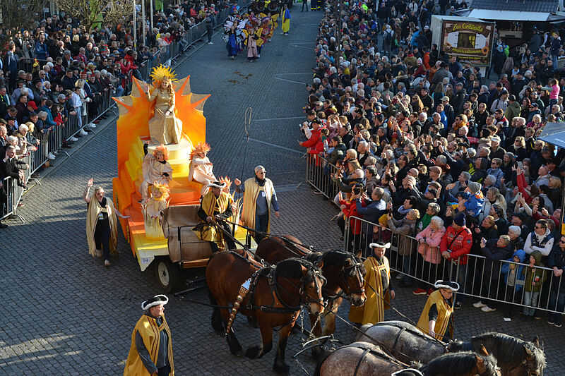 Ein Festwagen fährt durch eine Menschenmenge. Auf dem Wagen steht eine als Sonne verkleidete Frau.