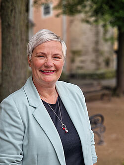 Ein Portrait. Eine Frau mittleren Alters schaut direkt in die Kamera und lächelt. Sie hat kurzes blondes Haar, trägt roten Lippenstift und ein schwarzes Oberteil