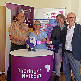 Drei Männer und eine Frau stehen vor Werbebannern. Drei der Personen stecken einen leuchtenden Stecker in eine lilafarbene Weltkugel als Symbol für den Glasfaseranschluss.