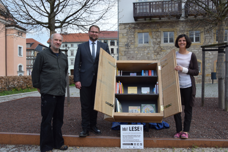 Drei Menschen stehen neben einer Holzkonstruktion. Dort können Bücher zum Verschenken hineingestellt werden. Die Konstruktion sieht aus wie ein Buch.