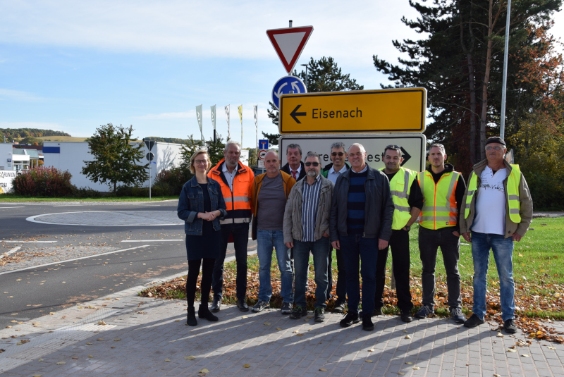 Vor einem Verkehrsschild steht eine Gruppe Menschen. Dahinter ist links im Bild ein Kreisverkehr zu sehen.