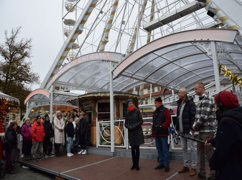 Eine Frau und drei Männer stehen vor einem Riesenrad. Links am Rand schauen Fahrgäste und Schüler gespannt.