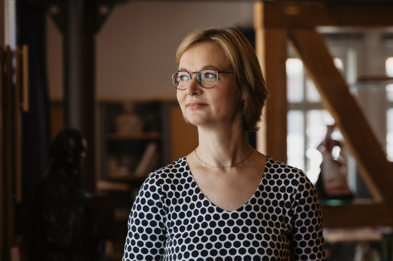 Eine Frau mit blonden Haaren und einem gepunkteten Oberteil blickt zur Seite. Im Hintergrund ist schemenhaft ein Büro erkennbar.