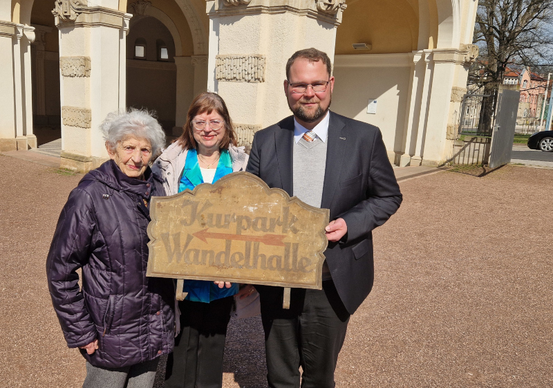 Eine alte Frau, ihre erwachsene Tochter und ein Mann im Anzug stehen vor einer historischen Halle. In den Händen halten sie ein altes Schild mit dem Hinweis "Kurpark" und "Wandelhalle".
