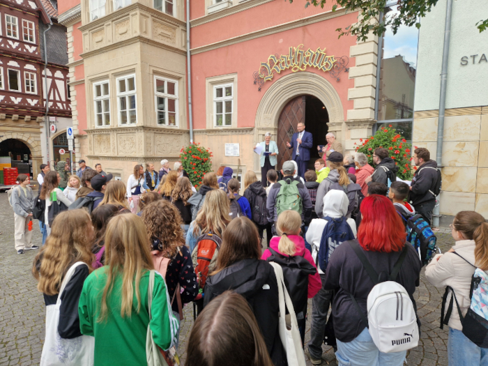 Auf den Stufen eines historischen Gebäudes stehen ein Mann und eine Frau. Vor ihnen stehen viele Jugendliche. Man sieht sie nur von hinten. Der Mann spricht in ein Mikrofon.
