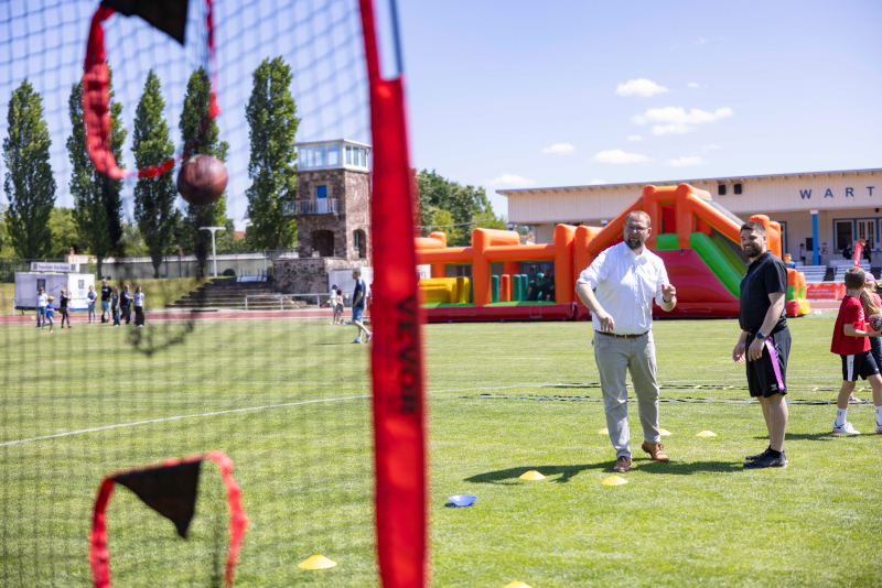 Ein Mann in einem weißen Hemd und ein sportlich gekleideter Mann stehen auf einem Rasen. Sie werfen einen kleinen Ball in ein Tor. Im Hintergrund ist eine Hüpfburg zu sehen.
