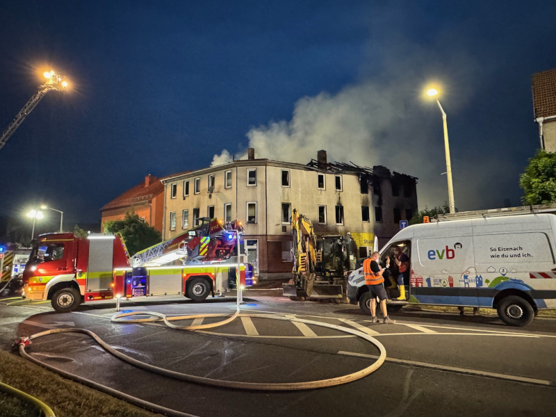 Mehrere Feuerwehrautos stehen im Dunkeln auf einer Straße. Rauch steigt aus einem Gebäude im Hintergrund auf.