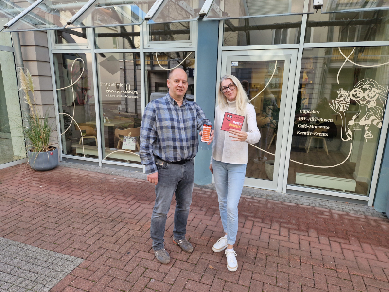 Eine Frau und ein Mann stehen vor einem Café. Sie halten einen Aufkleber und ein Handy in der Hand und zeigen es.