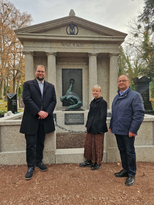 Zwei Männer und eine Frau stehen vor einem imposanten Portal. In der Mitte ist ein knieender Jüngling zu sehen, der an einer Türe zieht.