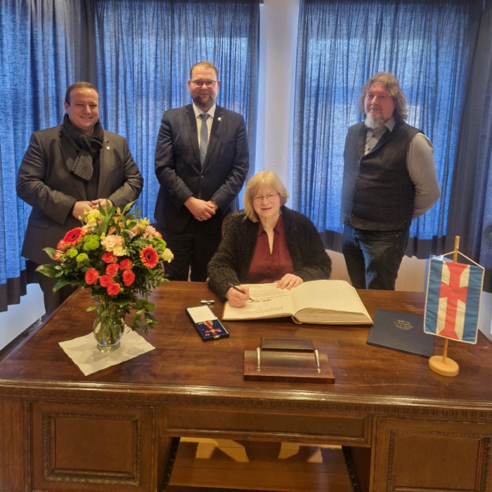 Eine Frau sitzt an einem Tisch und schreibt in ein großes Buch. Hinter ihr stehen drei Männer. Auf dem Tisch stehen ein Blumenstrauß und ein Fähnchen. Zudem ist ein Orden in einer Schatulle zu sehen.