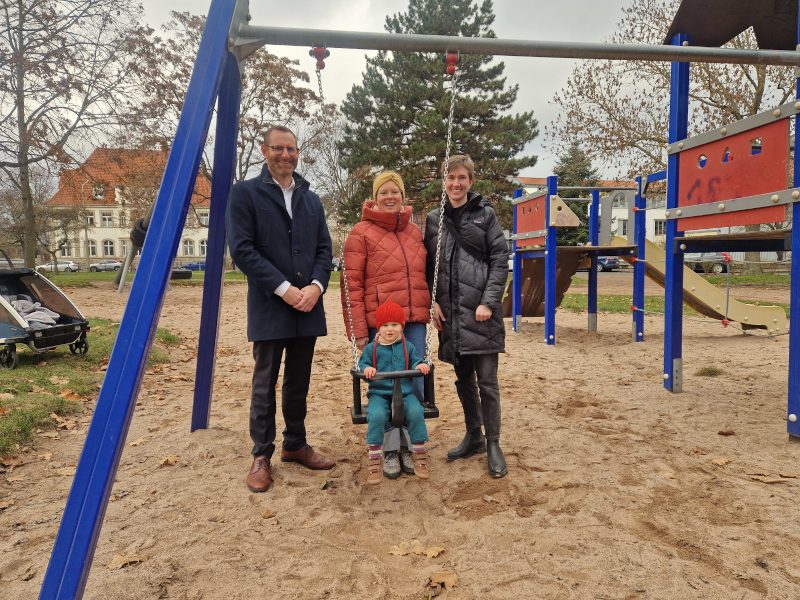 Zwei Frauen und ein Mann stehen an einer Schaukel, die für Kleinkinder geeignet ist. In der Schaukel sitzt ein Kind.