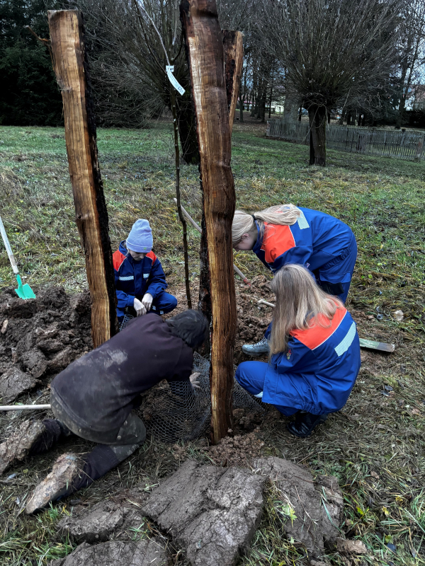 Vier Kinder, drei davon in Feuerwehruniformen, hocken auf einer Wiese und plflanzen einen Baum.