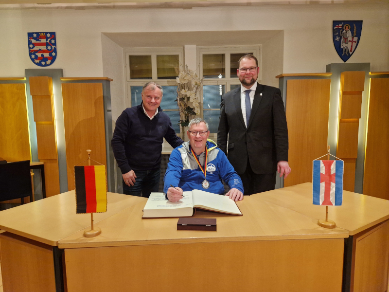 Ein Mann im Rollstuhl sitzt an einem Tisch und unterschreibt in einem großen Buch. Zwei Männer stehen links und rechts von ihm.