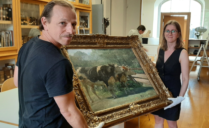 Ein Mann und eine Frau halten ein Gemälde. Das Bild zeigt Rinder und hat einen mächtigen goldenen Rahmen.