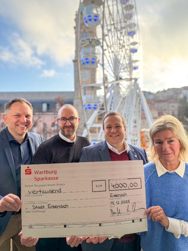 Drei Männer und einen Frau halten einen symbolischen Spendenscheck in den Händen. Darauf steht die Zahl 4000. Im Hintergrund ist ein Riesenrad zu sehen.