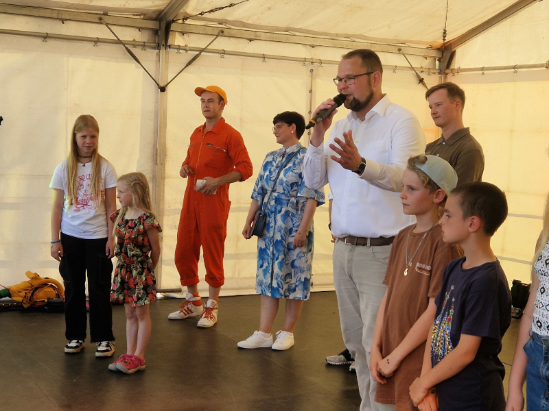 Ein Mann im weißen Hemd steht auf einer Bühne und spricht in ein Mikrofon. Neben ihm stehen Kinder.