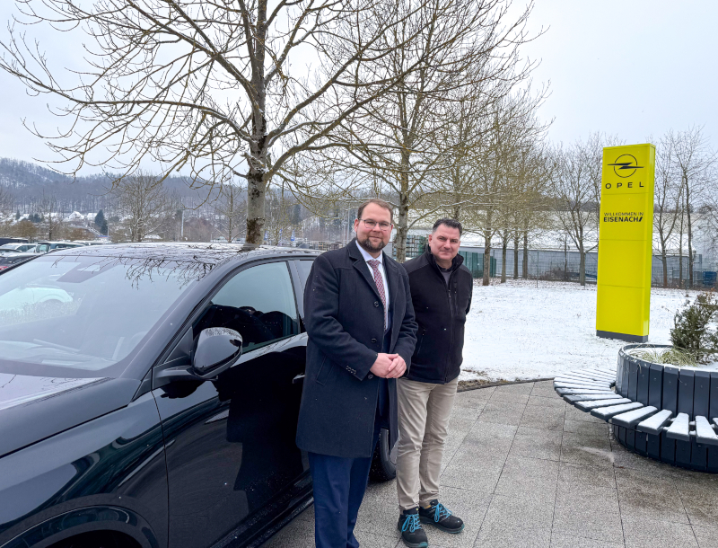 Zwei Männer stehen vor einem scharzen Auto. Im Hintergrund stehen Bäume und ein gelbes Schild.