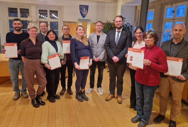 Fünf Frauen und sechs Männer stehen in einer Gruppe. Mehrere von ihnen halten Urkunden in der Hand. An der Wand hinter ihnen hängt ein Wappen.