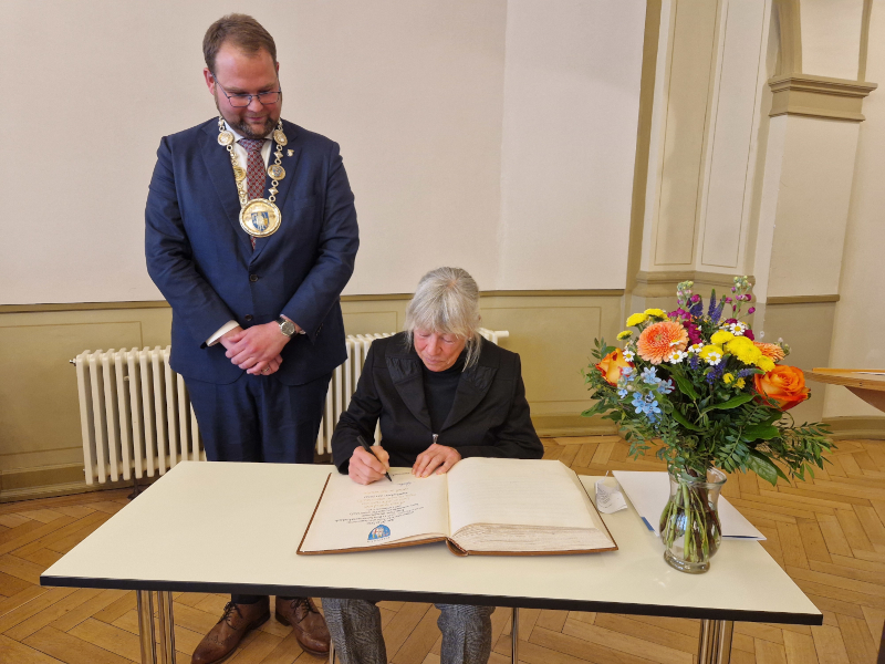Eine Frau sitzt an einem Tisch und schreibt in ein großes Buch. Links von ihr steht ein Mann im Anzug, der eine wuchtige Kette um den Hals trägt. Rechts auf dem Tisch Blumen.