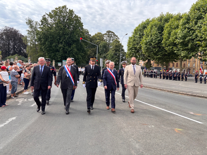 Mehrere Männer, teilweise mit Schärpen, laufen eine Straße entlang. Rechts und links stehen Menschen.