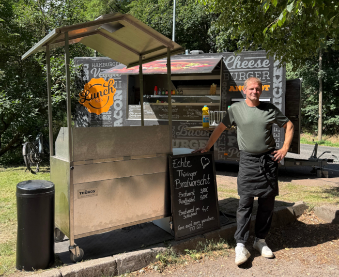 Ein Mann steht neben einem Grill. Hinter ihm ist ein Imbisswagen zu sehen.