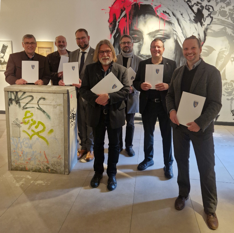 Sieben Männer mit Urkunden in den Händen stehen vor einem Wandbild (Graffiti).