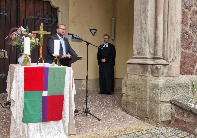 Ein Mann im Anzug und ein Mann im Talar stehen vor einem Portal. Links ist ein Altar mit Kreuz und Kerze aufgebaut.
