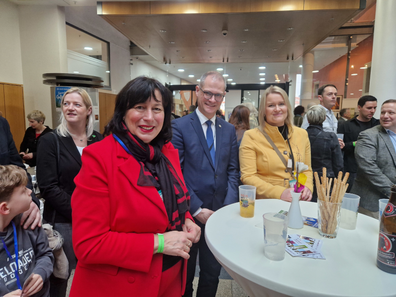 Zwei Frauen und ein Mann stehen an einem Stehtisch. Im Hintergrund Menschen
