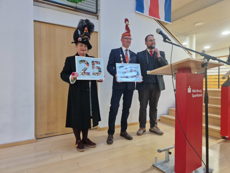 Eine Frau und ein Mann halten Bilderrahmen in den Händen. Daneben steht ein Mann im Anzug.