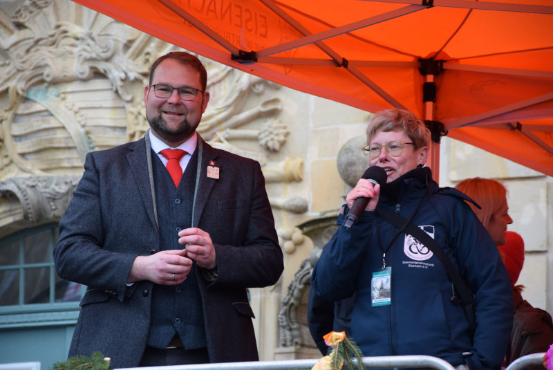 Ein Mann im Anzug und eine Frau mit einem Mikrofon in der Hand stehen auf einer Bühne.