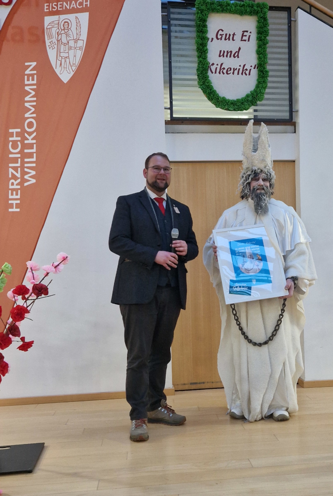 Ein als Winter verkleideter Mann und ein Mann im Anzug stehen nebeneinander. Der Winter hält einen Bilderrahmen in der Hand.