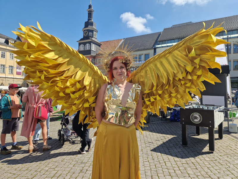 Eine junge Frau ist als goldener Engel verkleidet. Sie steht draußen auf einem Platz.