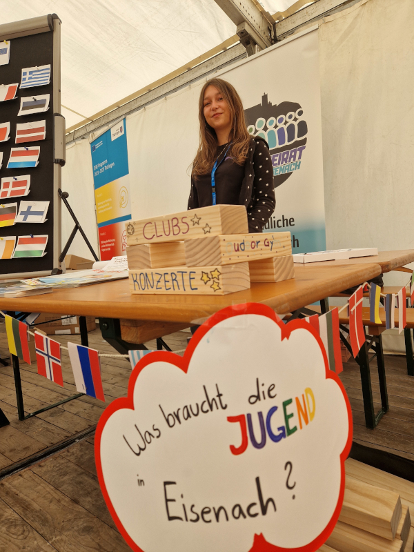 Eine junge Frau steht an einem Tisch, davor ein Schild.