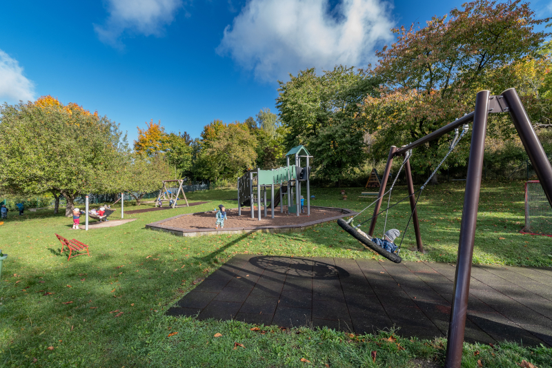 Grünes Außengelände mit Schaukeln, Klettertrum und spielenden Kindern