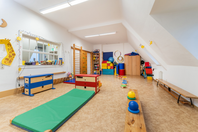 Spielzimmer eines Kindergartens mit Turnmatte, Regalen aus Holz, einer Bank an der Seite und Sprossenwand und Kletterwand