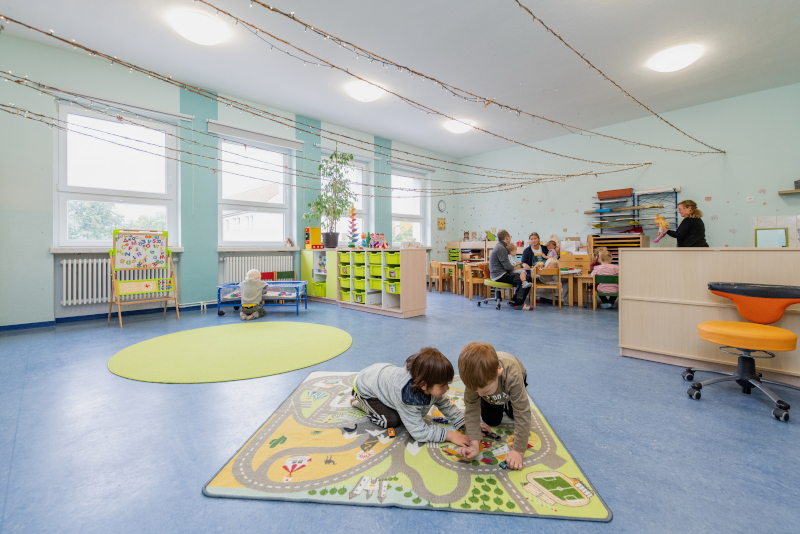 Großes Spielzimmer in einem Kindergarten mit Spieleteppich, auf dem zwei Kinder hocken.