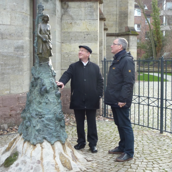 Zwei Männer in dunkler Kleidung stehen an einer Skulptur. Die Skulptur zeigt eine Frau, die auf einem Berg steht.