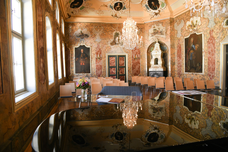 Ein festlicher Saal mit Kronleuchtern und Gemälden; links Fenster und vorn ein Flügel