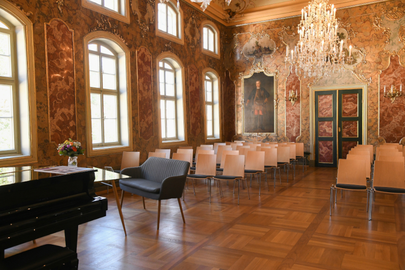 Saal mit Stühlen und Kronleuchter; Fenster links und Flügel sowie historischen Gemälden