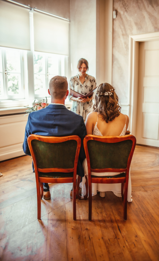 Ein Brautpaar sitzt auf Stühlen; Ansicht von hinten; Standesbeamtin steht im Hintergrund..