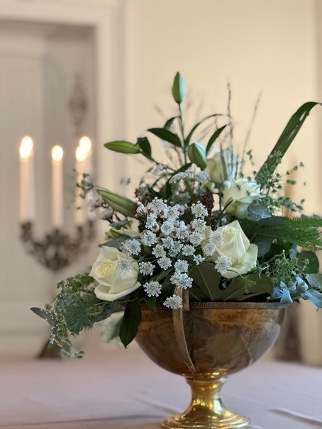 Ein Blumenarrangement in weiß in einer Schale; dahinter brennende Kerzen.