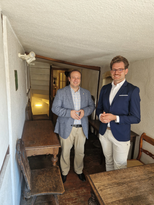 Zwei Männer im Jackett stehen in einem schmalen Raum. Wenige Holzmöbel sind erkennbar.