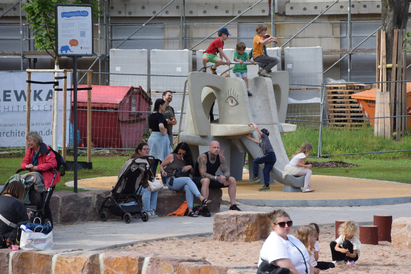 Erwachsene sitzen auf dem Spielplatz und Kinder rutschen auf der Elefantenrutsche.
